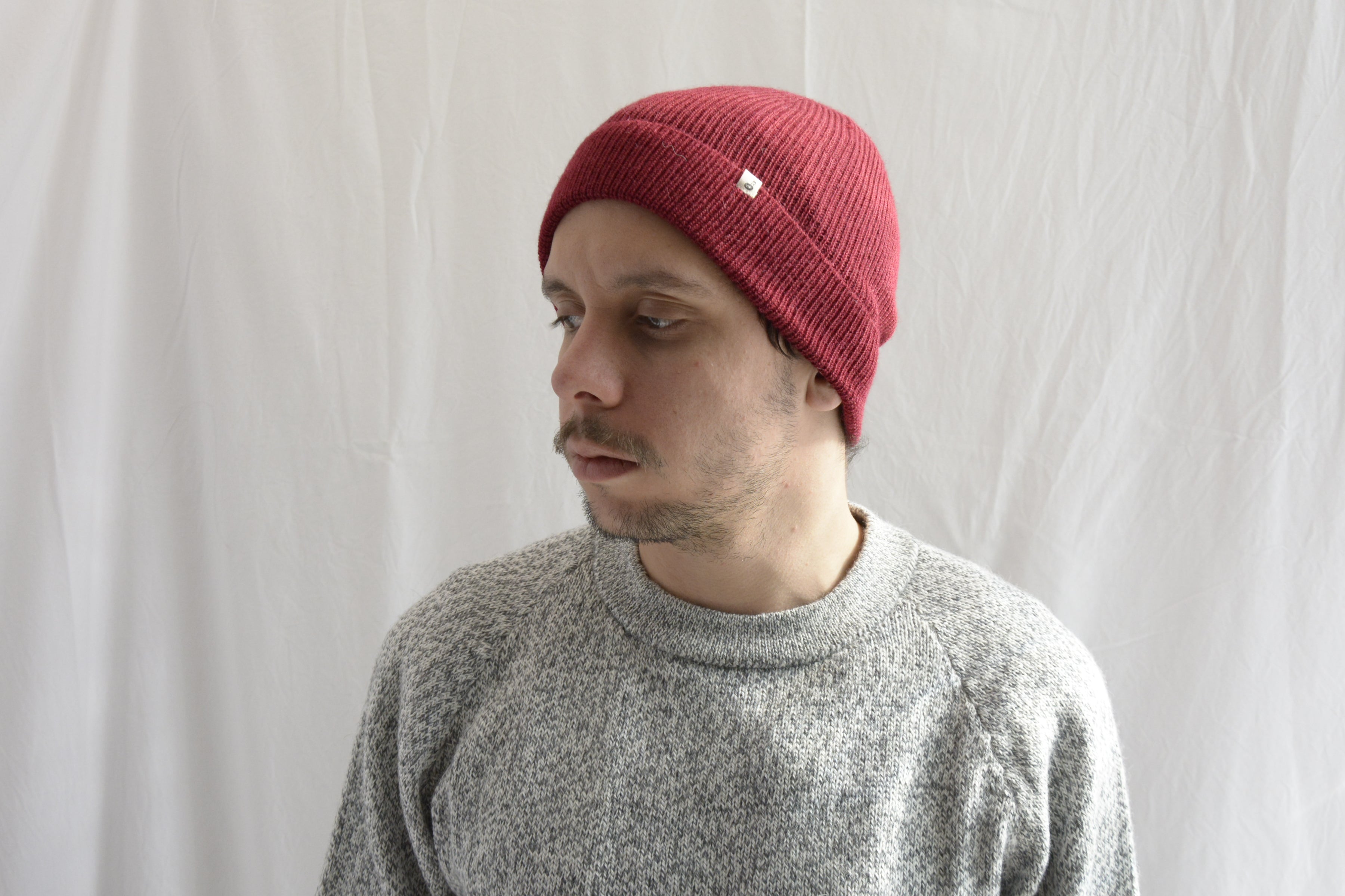 Man_wearing_brick_red_beanie_and_light_gray_sweater_alpaca_clothing_looking_down_beside